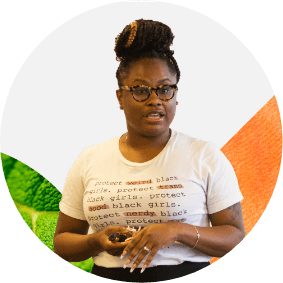 Teacher wearing glasses and a t-shirt with text, speaking while holding an object, against a colorful background.