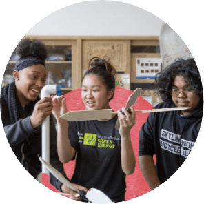 Three students collaborate on a project, assembling parts from a PVC pipe and cardboard in a classroom setting.